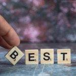 Hand places Scrabble tile “B” to complete the word “BEST,” symbolizing excellence, with a blurred colorful background.