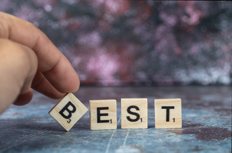 Hand places Scrabble tile “B” to complete the word “BEST,” symbolizing excellence, with a blurred colorful background.